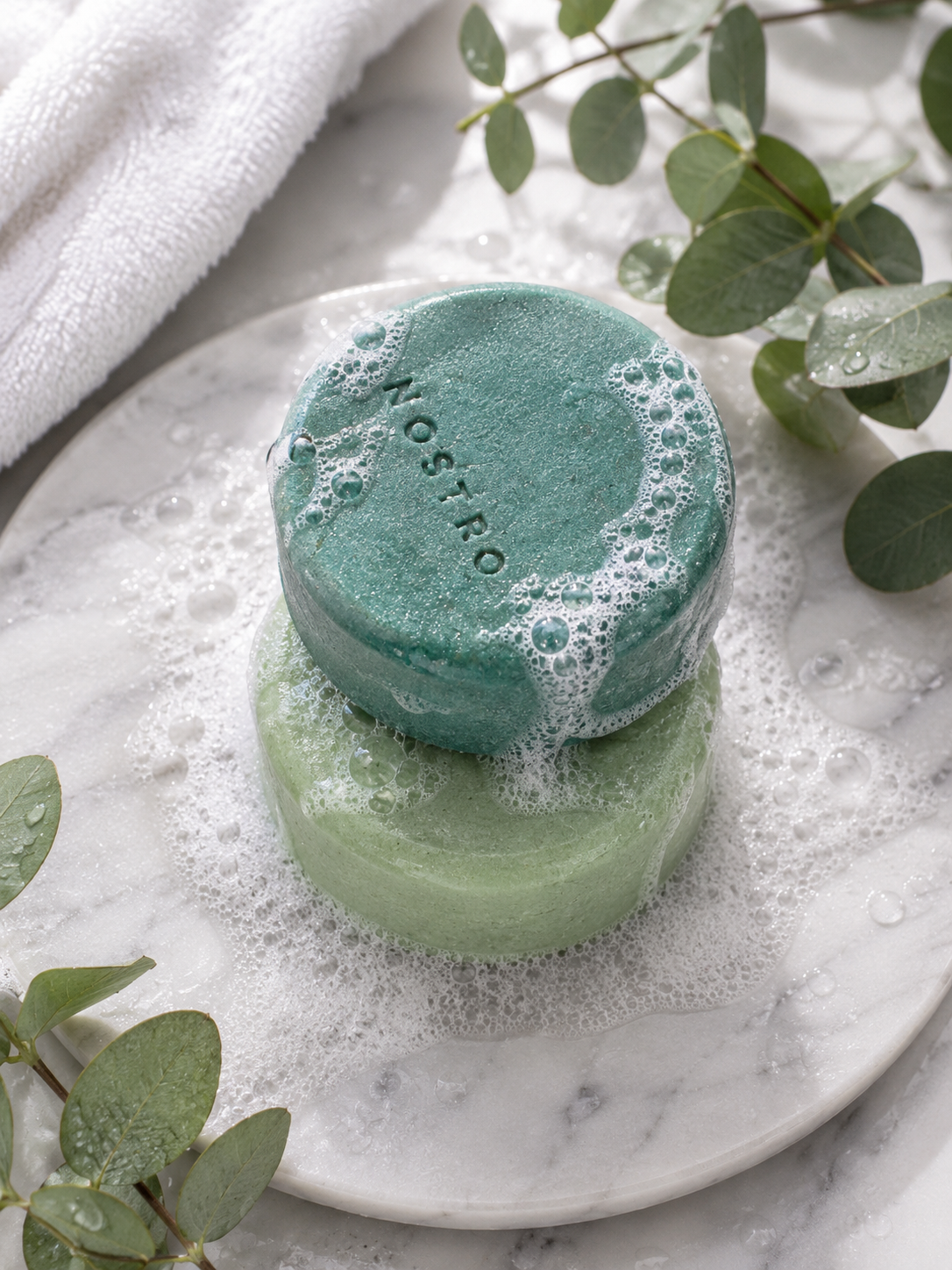 Barra de shampoo sólido Alpha azul verdoso con espuma sobre mármol blanco y eucalipto